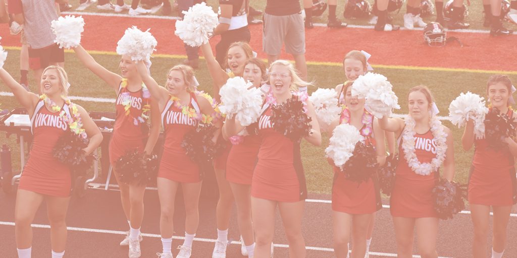 smiling cheerleaders on the sidelines of a field, laughing and shaking pom poms