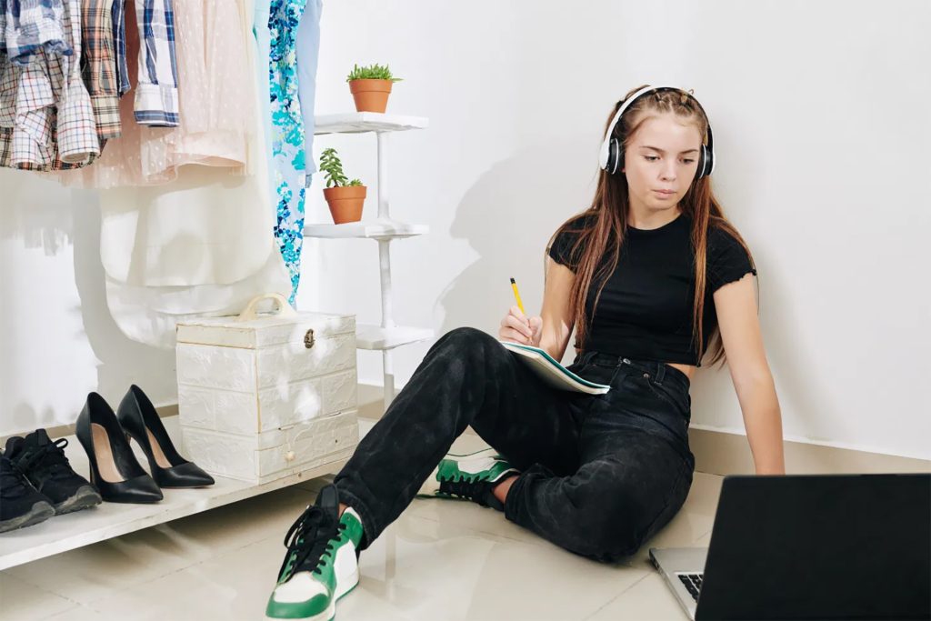 a teen wearing headphones working on homework, sitting on the floor with a laptop